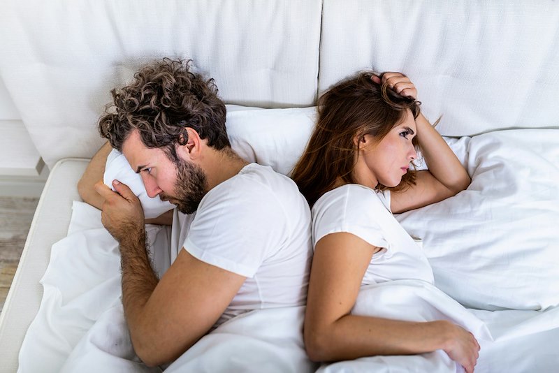 A husband and wife lay in bed opposite each other, upset