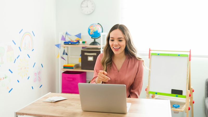 A teacher sitting at a table in a classroom points at her laptop.