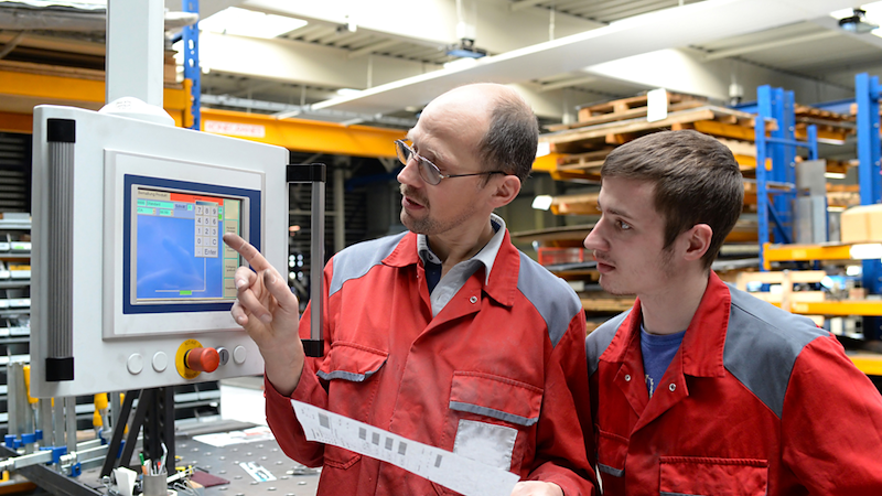 Two men in red work uniforms examine a machine control screen inside a factory.