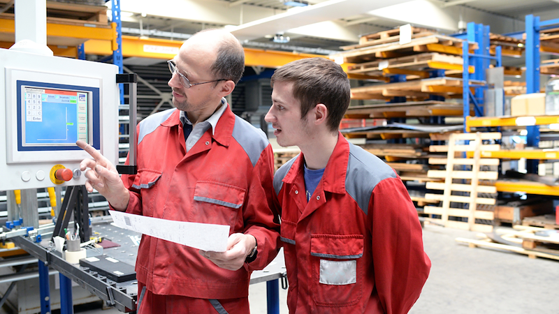 Older worker pointing out machine controls to a younger coworker