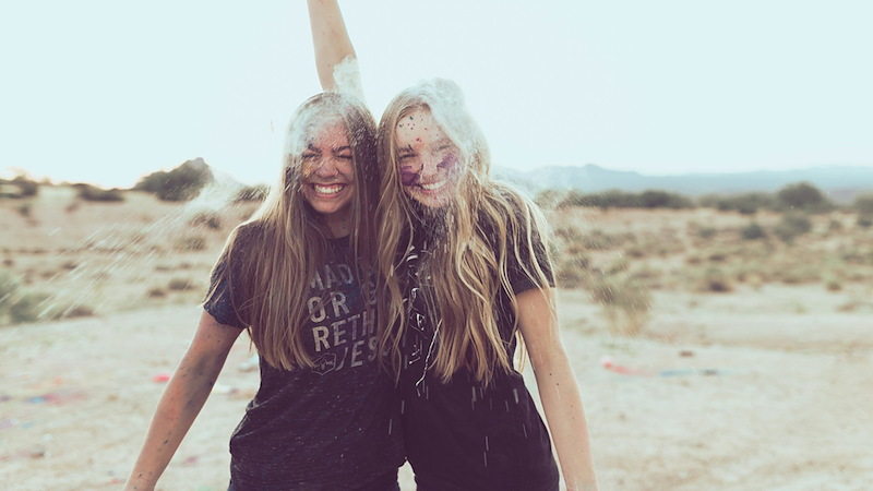 Two teenage girls smile together at the beach, while one throws sand over their heads.