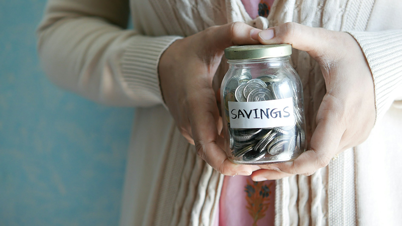 Close-up of a woman holding a jar full of silver coins with a label that says ‘savings’.