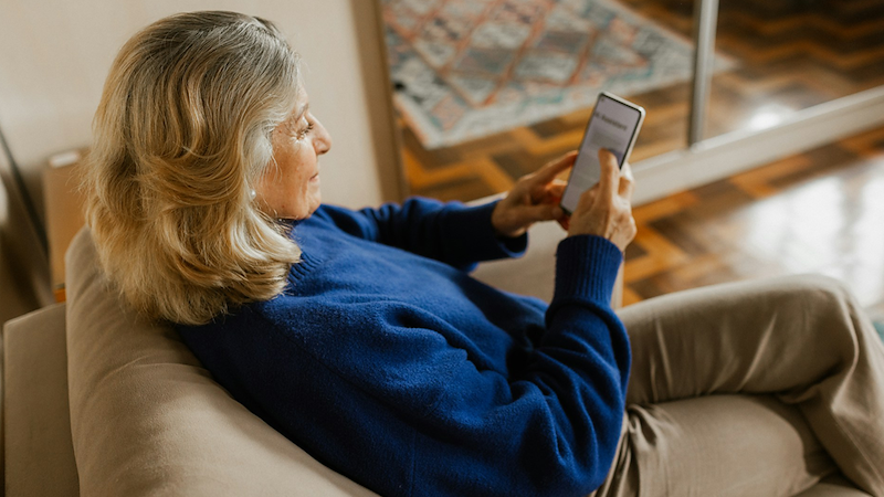 An older woman sits on a couch with crossed legs and looks at her phone.