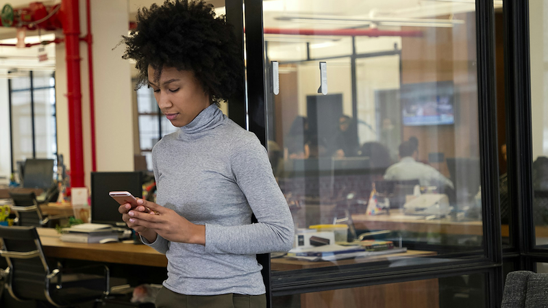 An employee leans against the glass door of an office, checking her phone.