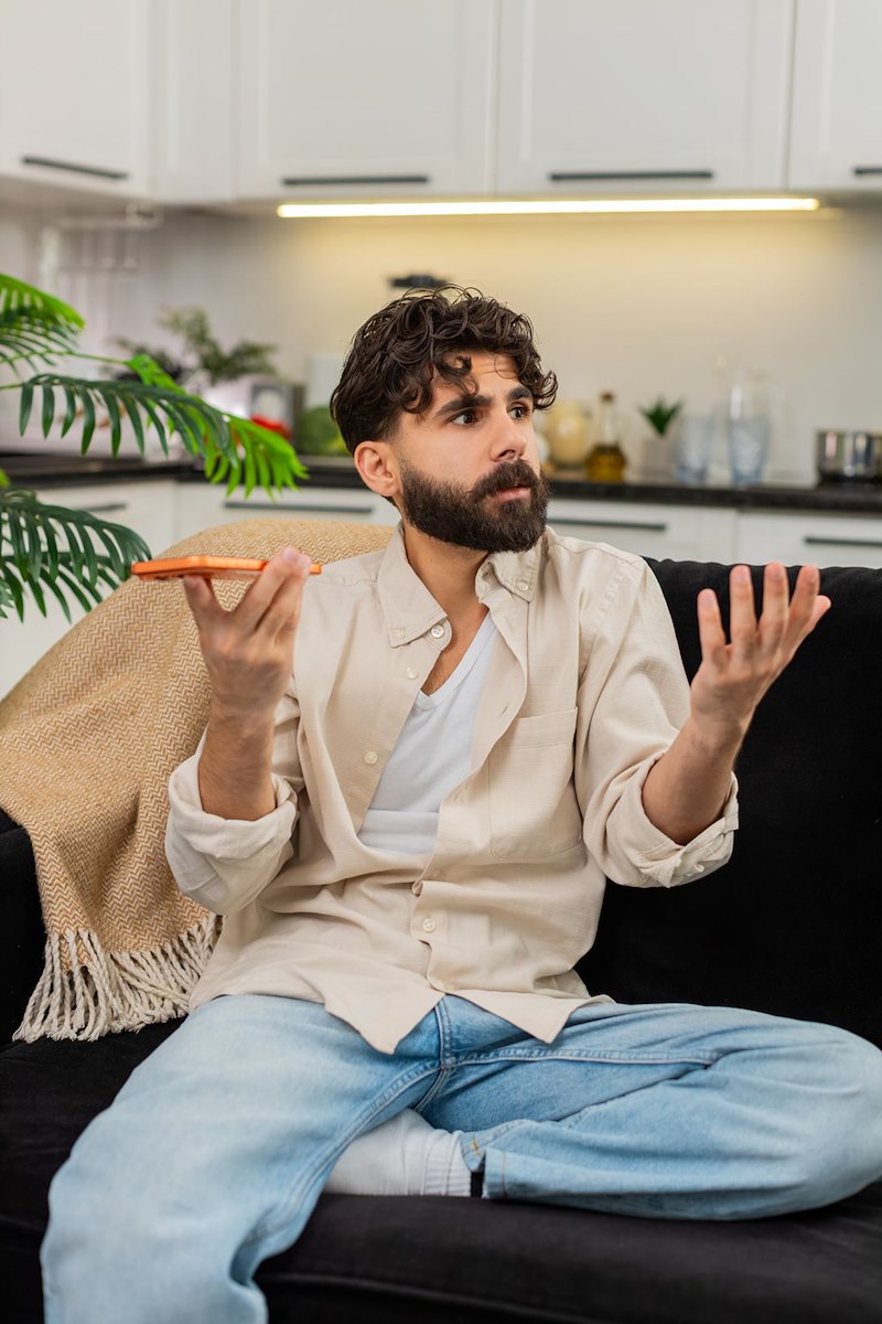 Bearded man sits cross-legged on a couch holding a phone and gesturing with both hands.