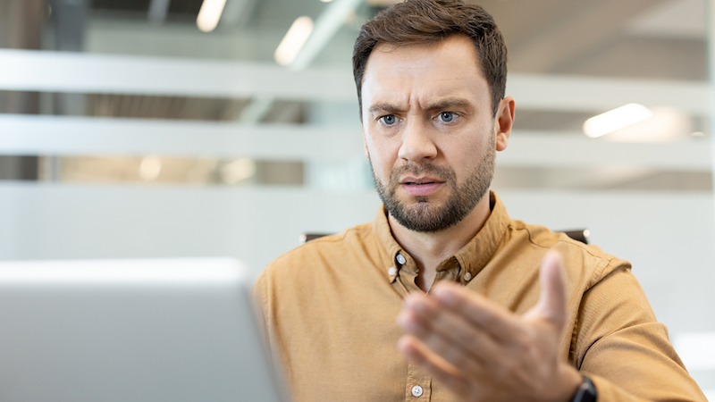 Man looks confused at a laptop while raising one hand in a questioning gesture.