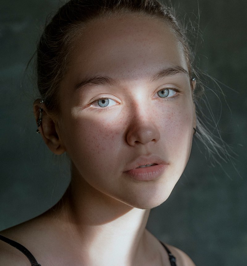 Close-up portrait of a young woman with blue eyes and a subdued expression against a dark background.