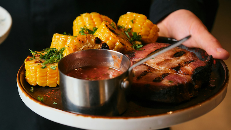 The hand of a waiter carrying a plate with steak, sauce, and sweetcorn on it.