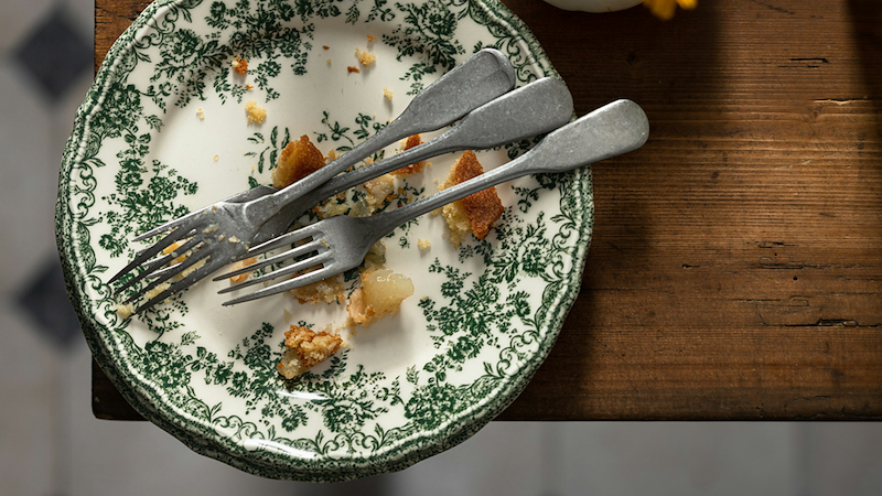 A plate sits on the edge of a table with crumbs and several forks on it.
