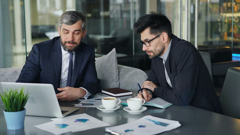 A boss and employee look at a laptop on a table with papers and coffee cups.