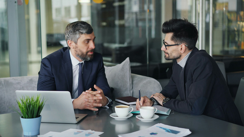 A boss and employee discuss something at a table with papers, a laptop, and coffee cups.