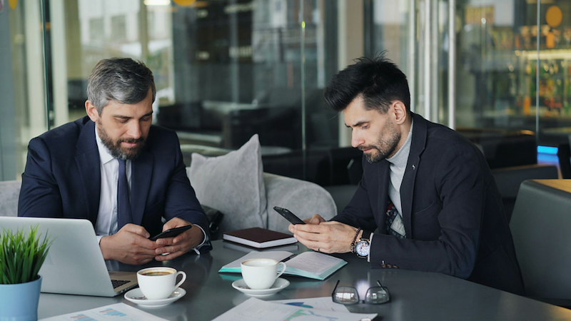 A boss and an employee sit at a table, looking at their phones.