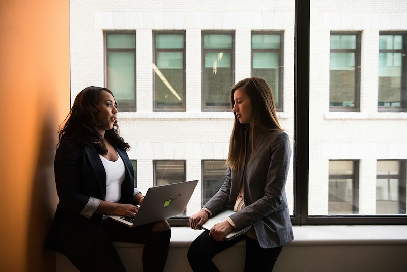 Two female coworkers discuss something by a window
