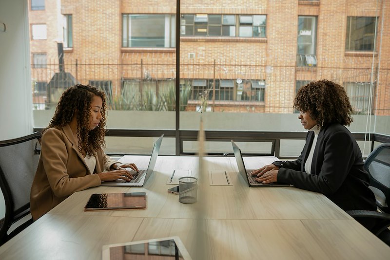 Two coworkers sit at a table with laptops