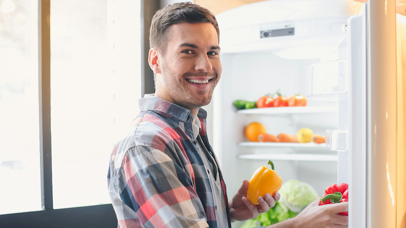 A man smiles as he puts bell peppers in a refrigerator.