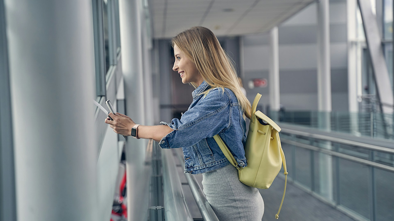 A woman smiles as she looks out the window of an air bridge and holds her phone.
