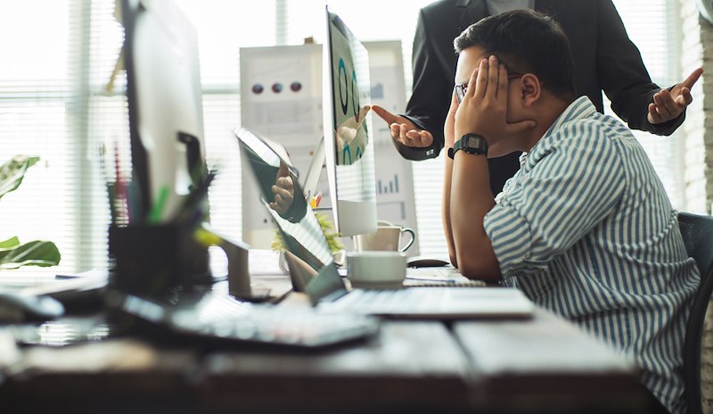 A frustrated office worker sits at his desk while a colleague speaks behind him.