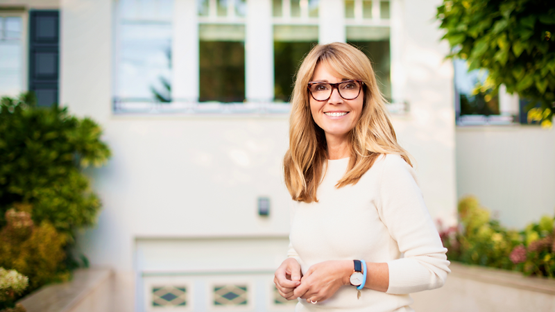 Smiling woman with glasses stands outside a large white house surrounded by greenery.