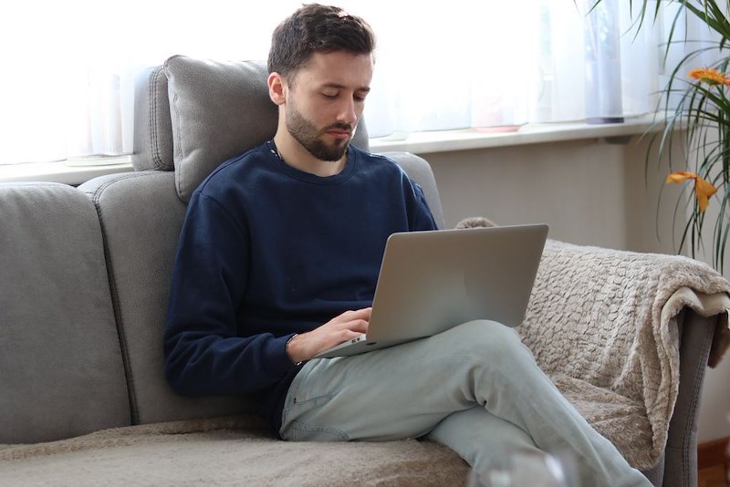 Man sits on a couch using a laptop at home, wearing a dark sweater and light pants.