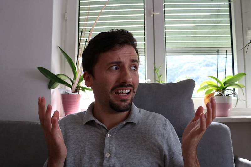 Man sits indoors with both hands raised and a confused expression