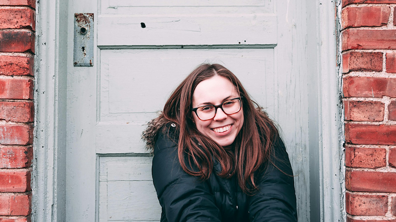A woman wearing a coat smiles as she sits in front of a door on a brick wall.