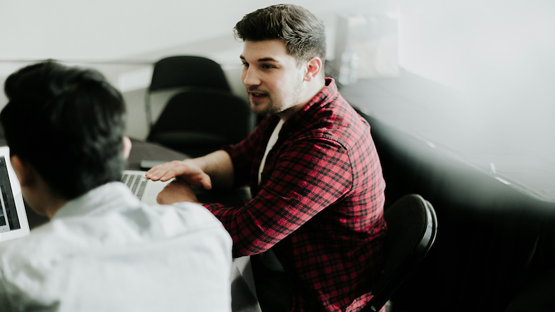 A man sitting by another man speaks, with their laptops on the table in front of them.