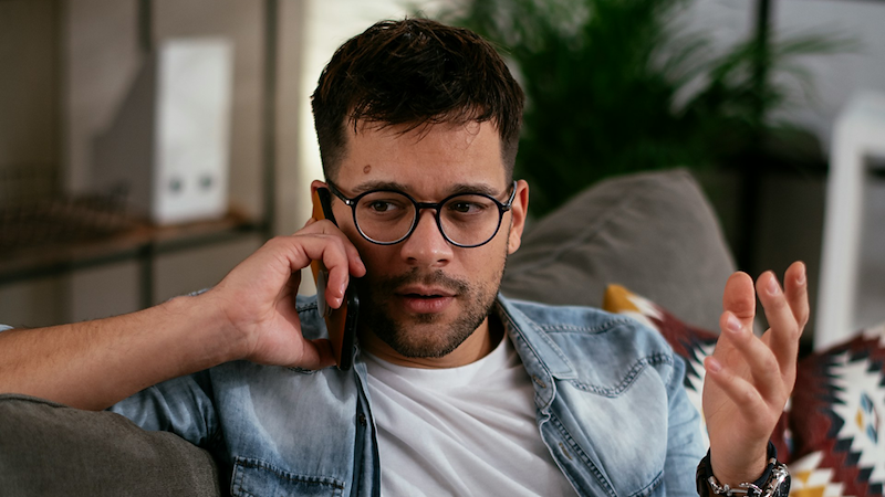 A man gestures while sitting on a couch and taking a phone call.
