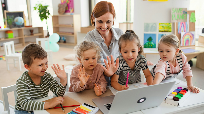 A female daycare assistant and four children sitting at a table wave at a laptop.