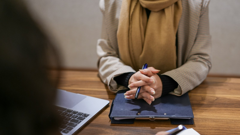 Close-up of an interviewer clasping her hands above a clipboard with a laptop beside her.
