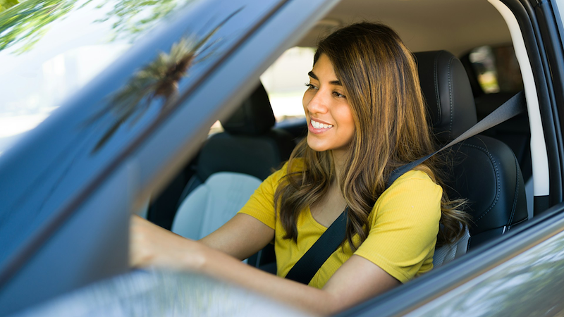 A woman smiles while driving a car with the window rolled down.
