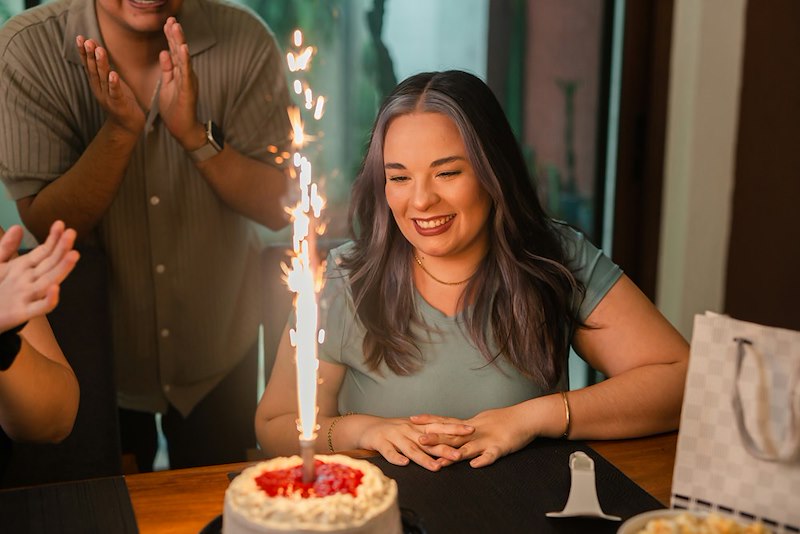 A woman sits in front of a birthday cake with a sparkler