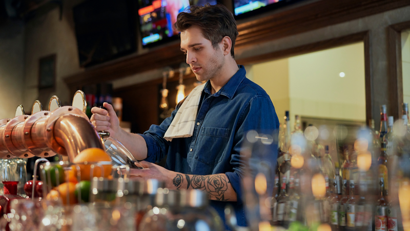Bartender in a blue shirt works behind a bar with beer taps, bottles, and glasses around him.