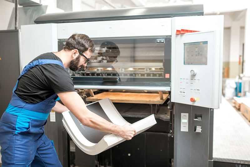 Print shop employee handling a stack of printed material beside an industrial printing machine.