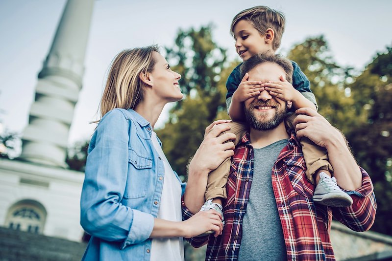 A happy mother and father walk outdoors, the son smiles atop his father's shoulders while he covers his dad's eyes.