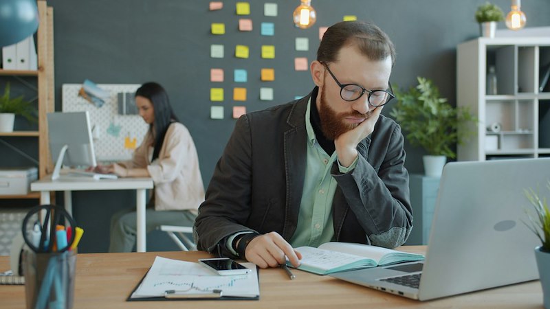 A man sits bored while tied to his desk working due to his boss's demands