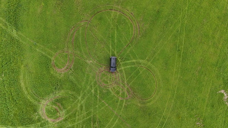 Aerial view of a car surrounded by looping tire tracks across a large green grassy field.