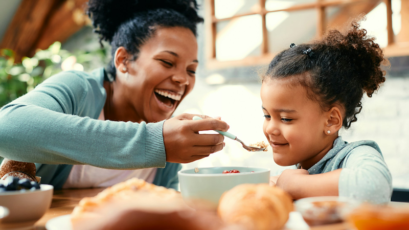 A mom laughs as she feeds her young daughter breakfast at the table.