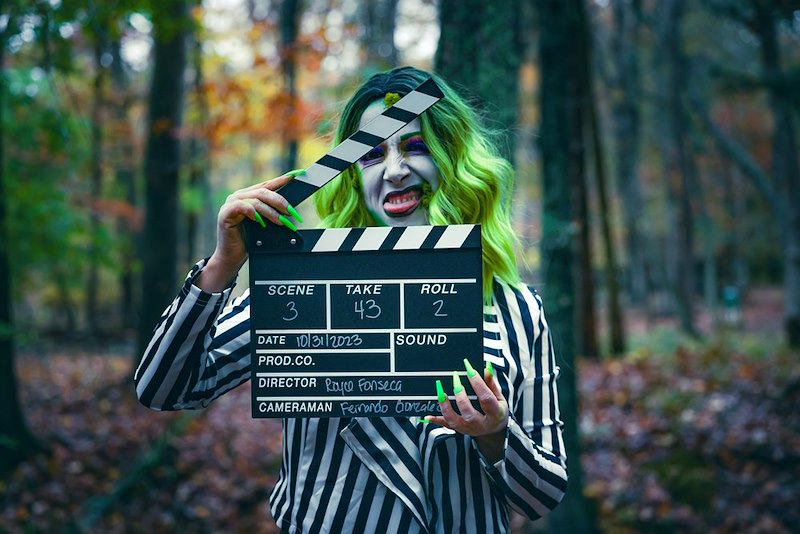 A person in a villain costume holds a clapperboard in a forest