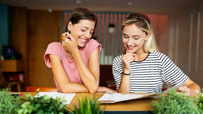 Two female friends smile as they sit at a table in a restaurant, looking at a menu.