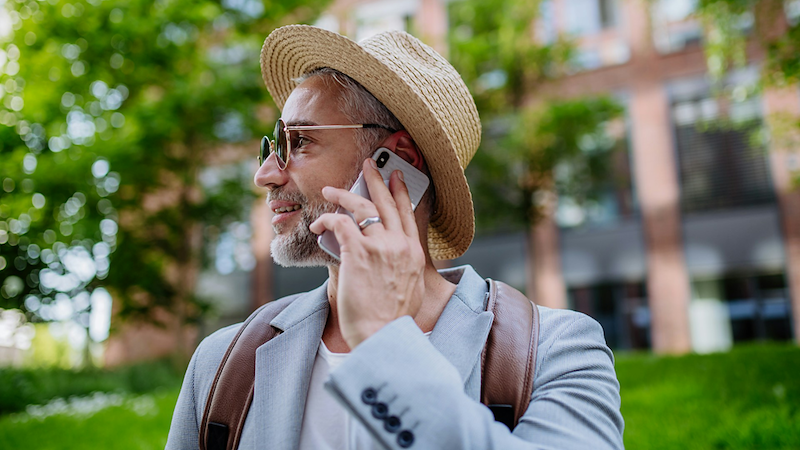 A businessman takes a call in a park, wearing a straw hat and sunglasses.