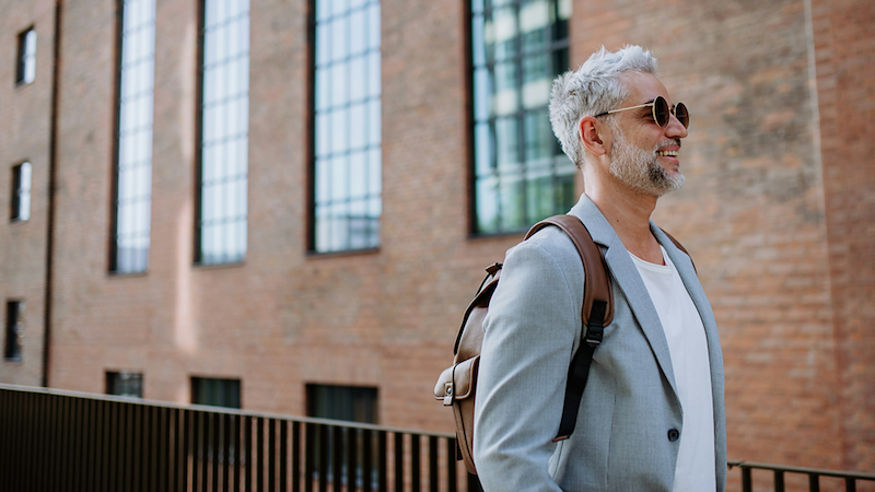 A businessman walks past a building, wearing sunglasses and a backpack.