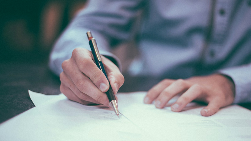 A close-up of a man in a shirt writing on some paper.