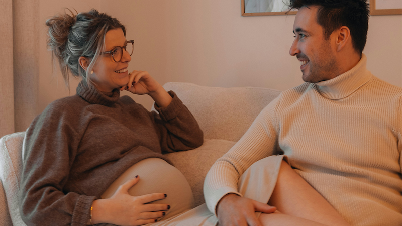 An expecting mom puts her legs on her partner's lap as they sit on the couch together.
