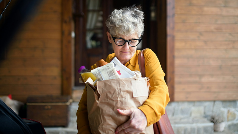 A woman carries a full paper bag of groceries next to the open trunk of a car.