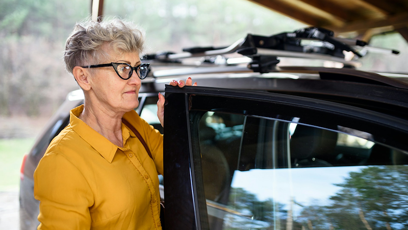 An older woman places her hand on top of the open door to the driver's seat of a car.