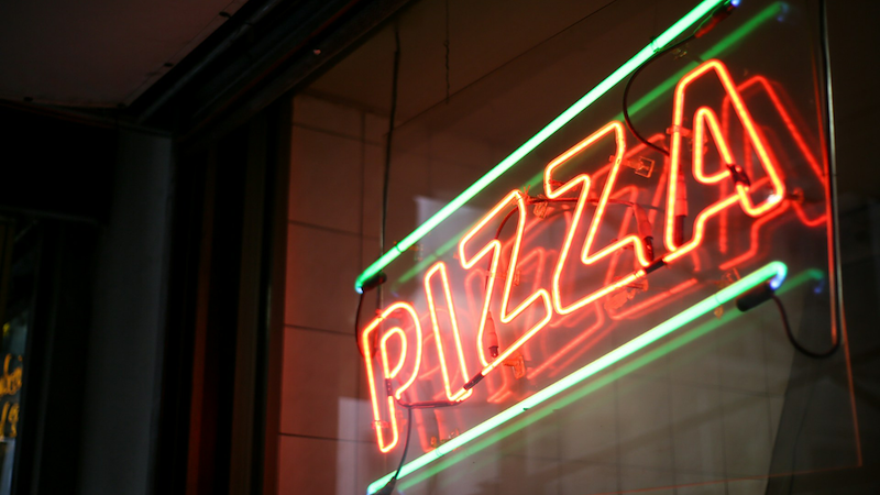 A red and green neon sign in a restaurant window that says ‘PIZZA’.