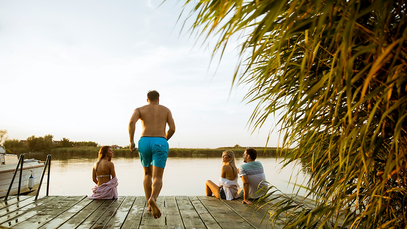 A man in swimwear runs along a lakeside dock as his friends sit at the end, watching him.