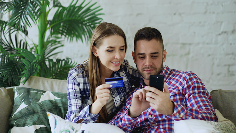 A couple sitting on a couch together, the man holding his phone and the woman holding a credit card.