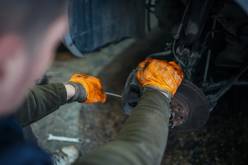 A mechanic wearing brown leather work gloves and a green workers over shirt attaches a brake caliper with a wrench.