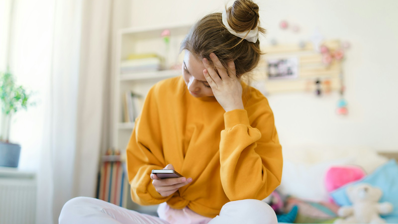 A teenage girl sitting cross-legged on a bed puts her head in her hand while looking at her phone.